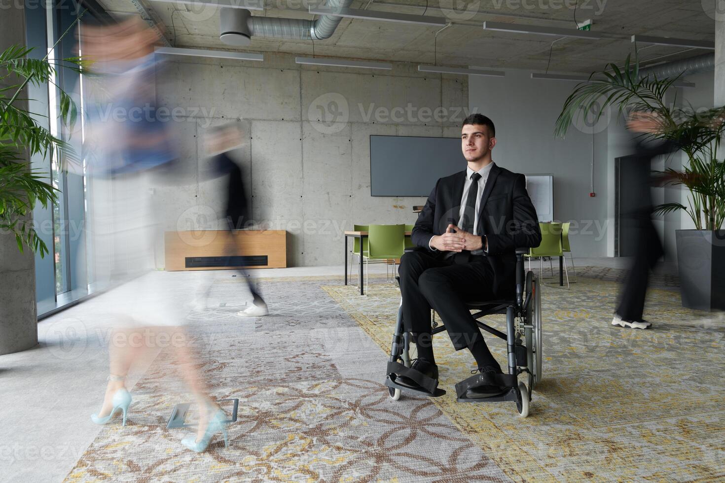 A businessman in a wheelchair navigating through a busy office, surrounded by his colleagues who are actively engaged in their work and collaboration photo