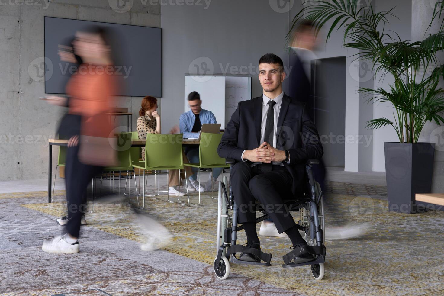 A businessman in a wheelchair navigating through a busy office, surrounded by his colleagues who are actively engaged in their work and collaboration photo