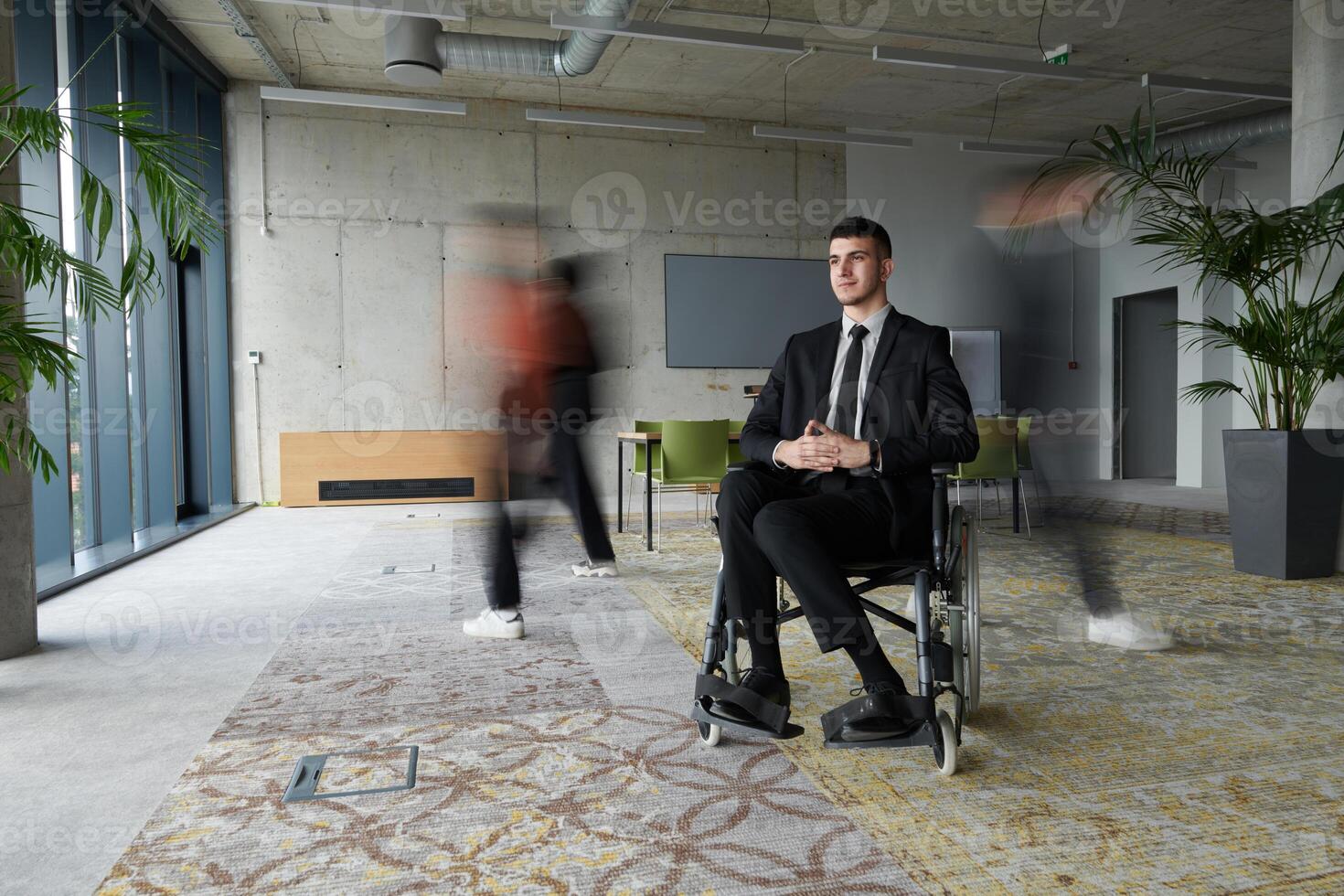 A businessman in a wheelchair navigating through a busy office, surrounded by his colleagues who are actively engaged in their work and collaboration photo