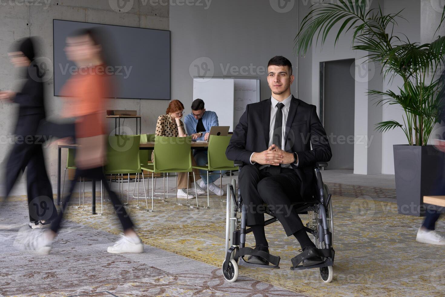 A businessman in a wheelchair navigating through a busy office, surrounded by his colleagues who are actively engaged in their work and collaboration photo