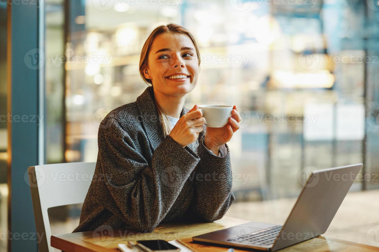 Stylish female freelancer is drinking coffee in cafe while working on laptop near window photo