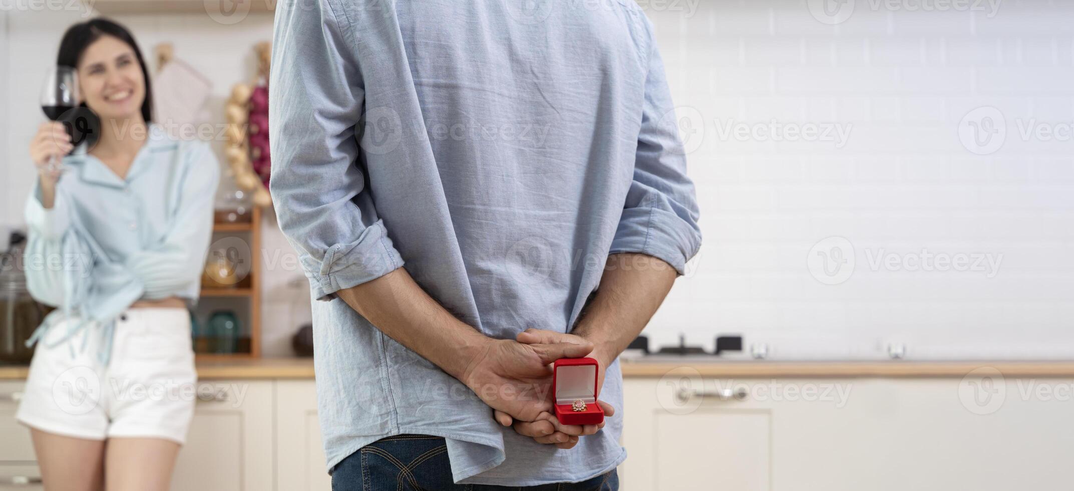 Multiethnic couple having romantic dinner in home kitchen. A man stands with a box with a wedding ring behind him. photo