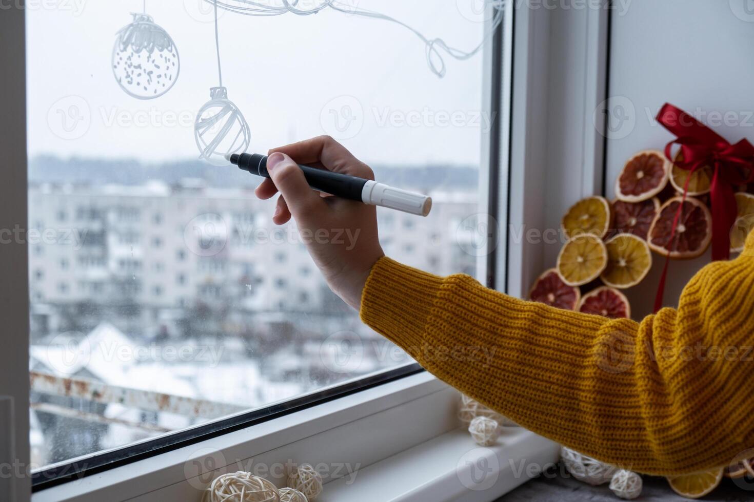 Creative enthusiastic unrecognizable woman in sweater paint window with white marker. Advent candle decoration on big clear window. Snowy winter outside. Holiday preparation photo