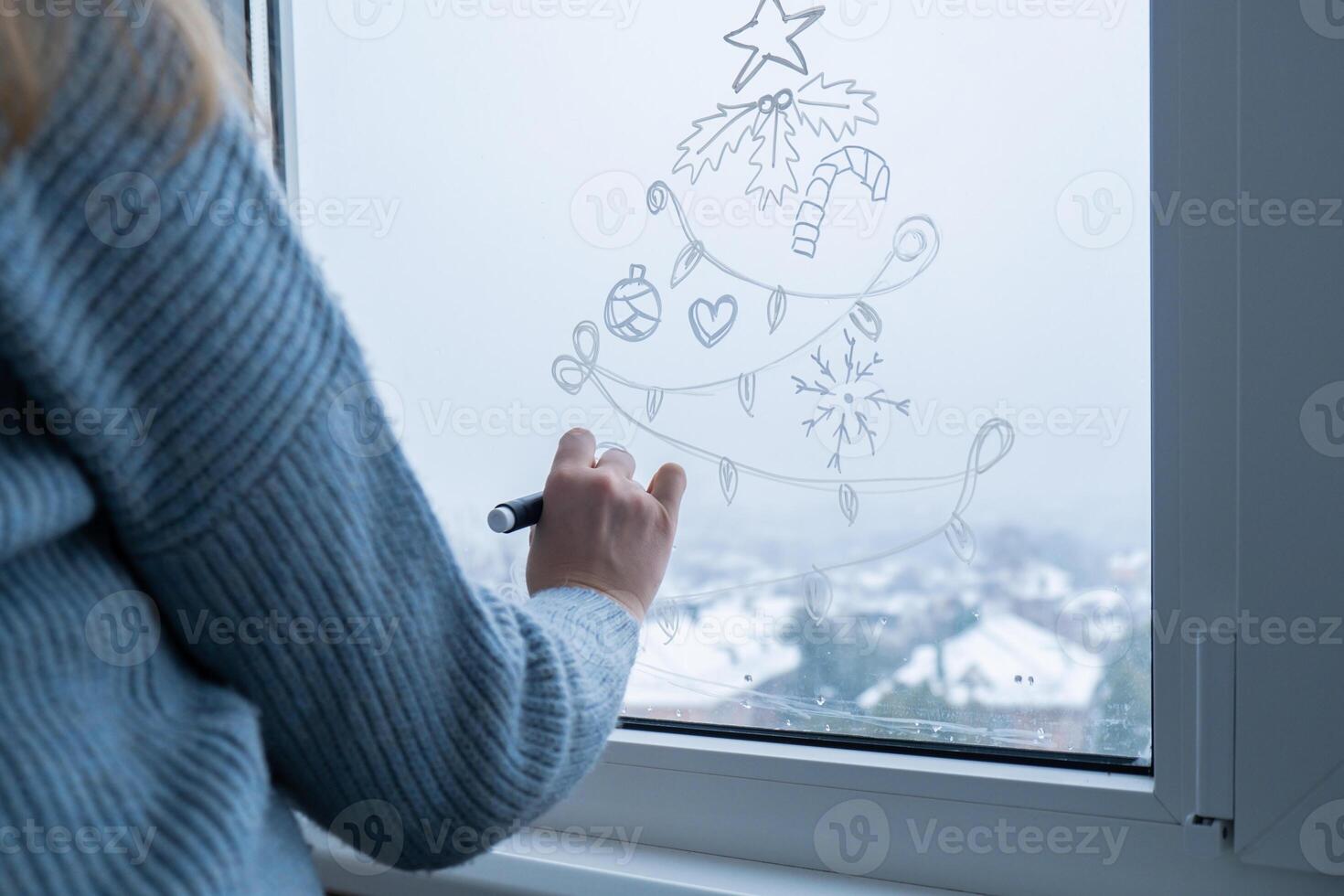 Creative enthusiastic unrecognizable woman in sweater paint window with white marker. Christmas tree decoration on big clear window. Snowy winter outside. Holiday preparation photo
