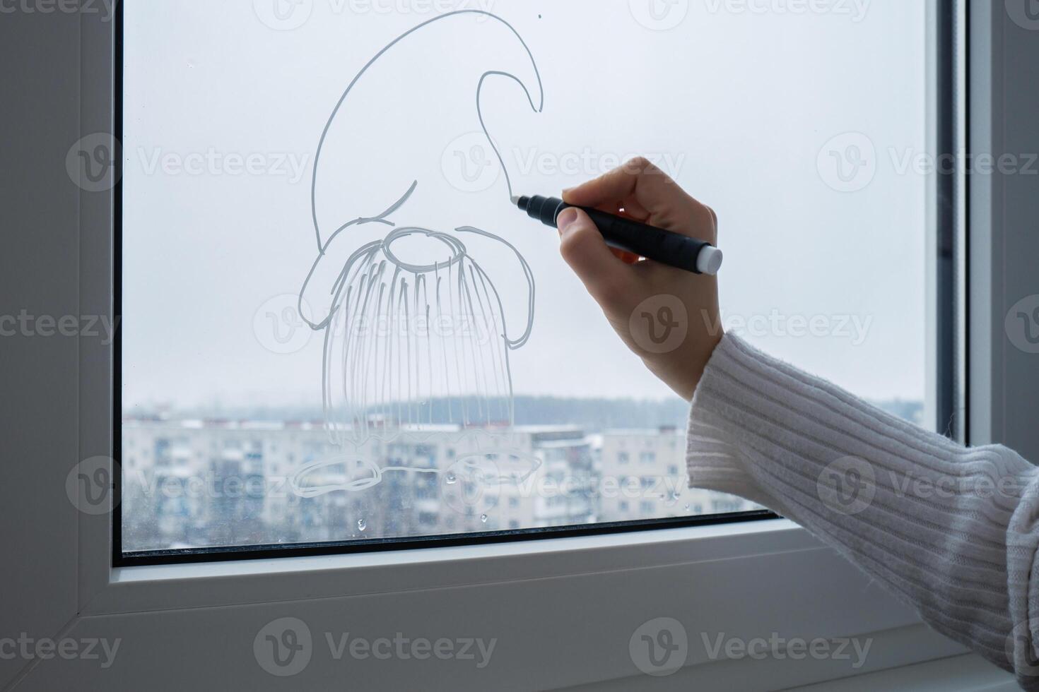 Creative enthusiastic unrecognizable woman in sweater paint window with white marker. Festive gnomes decoration on big clear window. Snowy winter outside. Holiday preparation photo