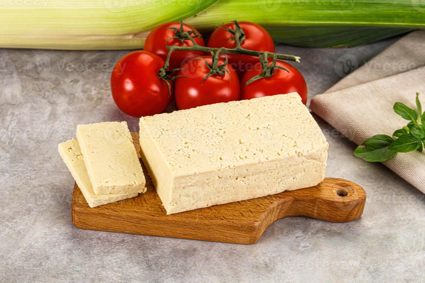 A piece of tofu on a cutting board next to a tomato and a piece of corn photo