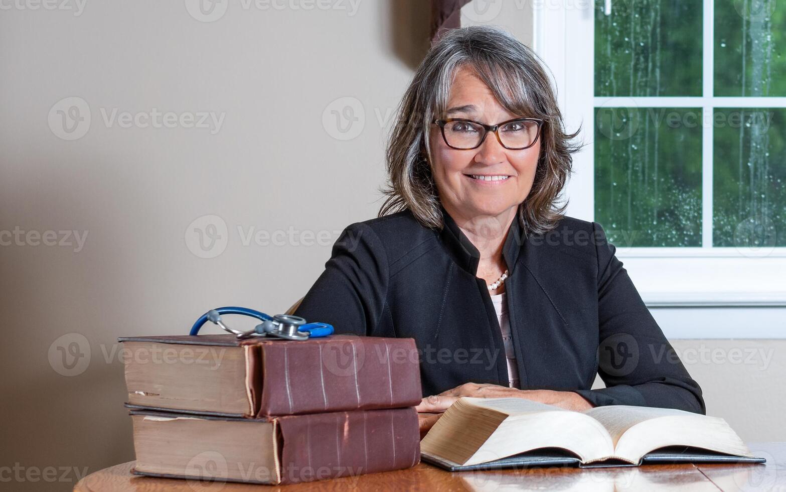 Female doctor sitting at a desk wearing glasses and a black coat with a stack of books and a stethoscope in the foreground, an open book in front of her and a window in the background. photo