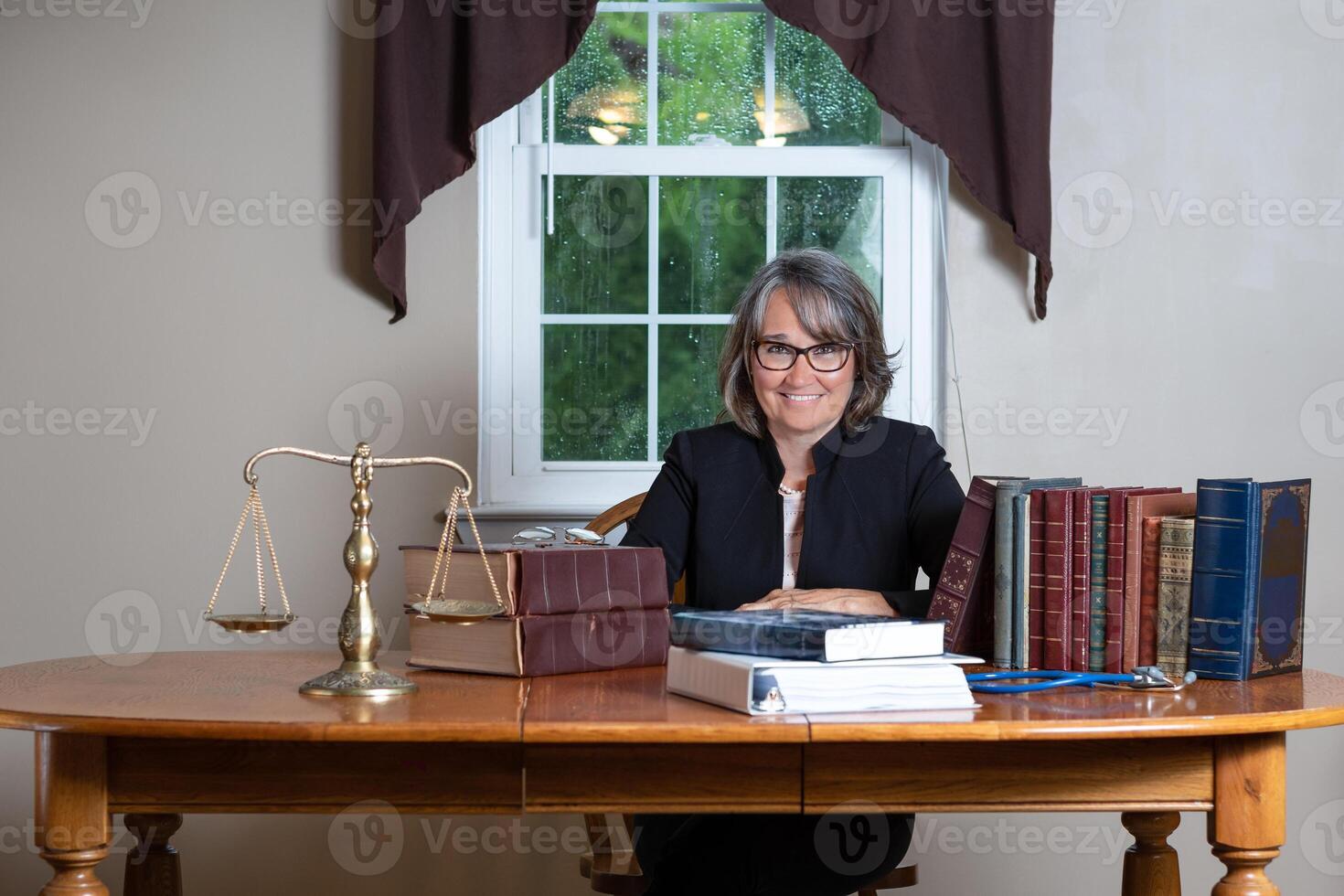 Woman attorney wearing glasses and a black coat sitting at a table behind stacks of legal books, a balance scale, and a stethoscope in front of a window smiling. photo