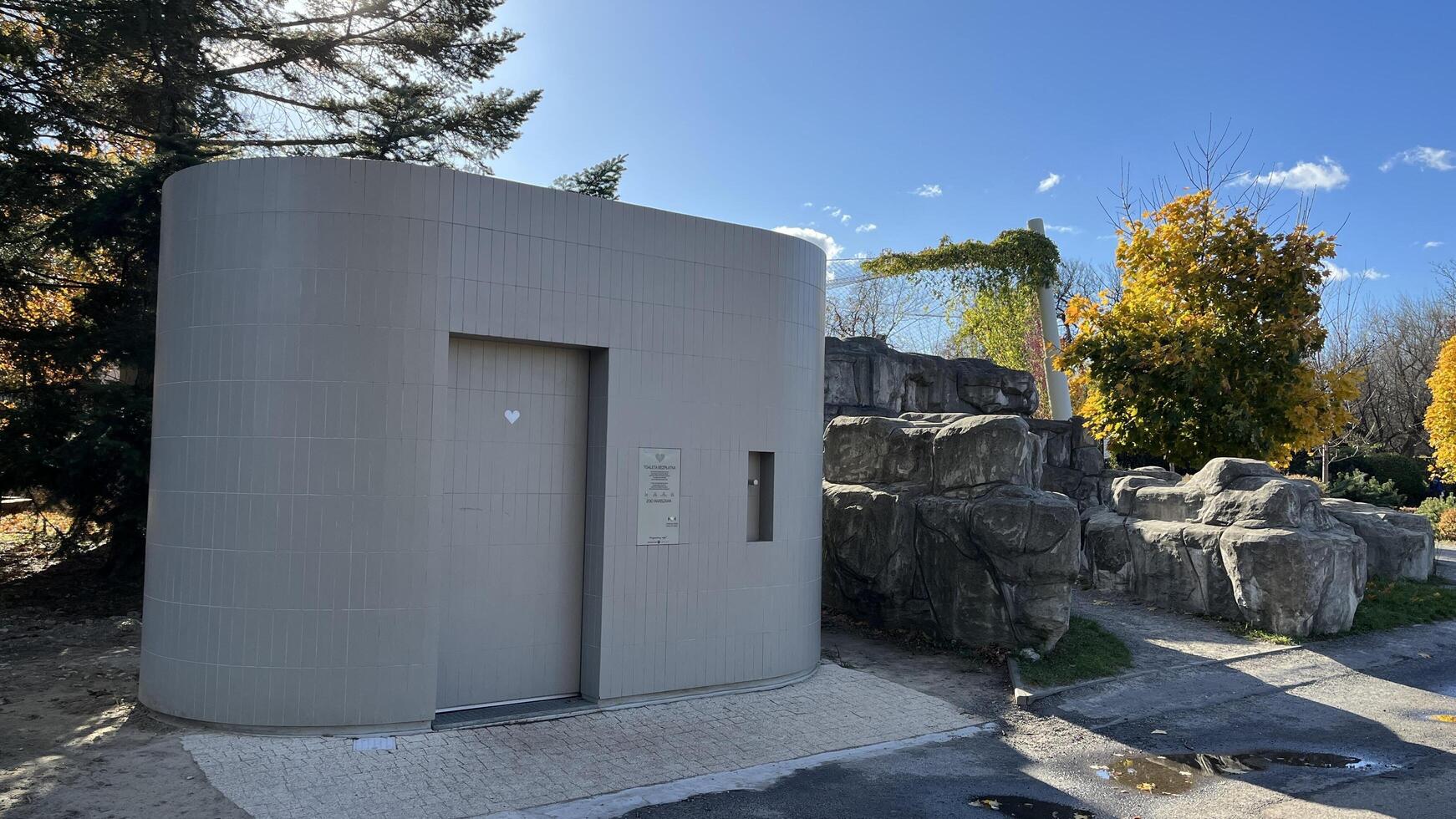Warsaw, Poland. November 02, 2024. A modern, curved toilet building with a gray exterior. It has a single door and a small window. Behind the building, there is a rock formation covered in greenery photo
