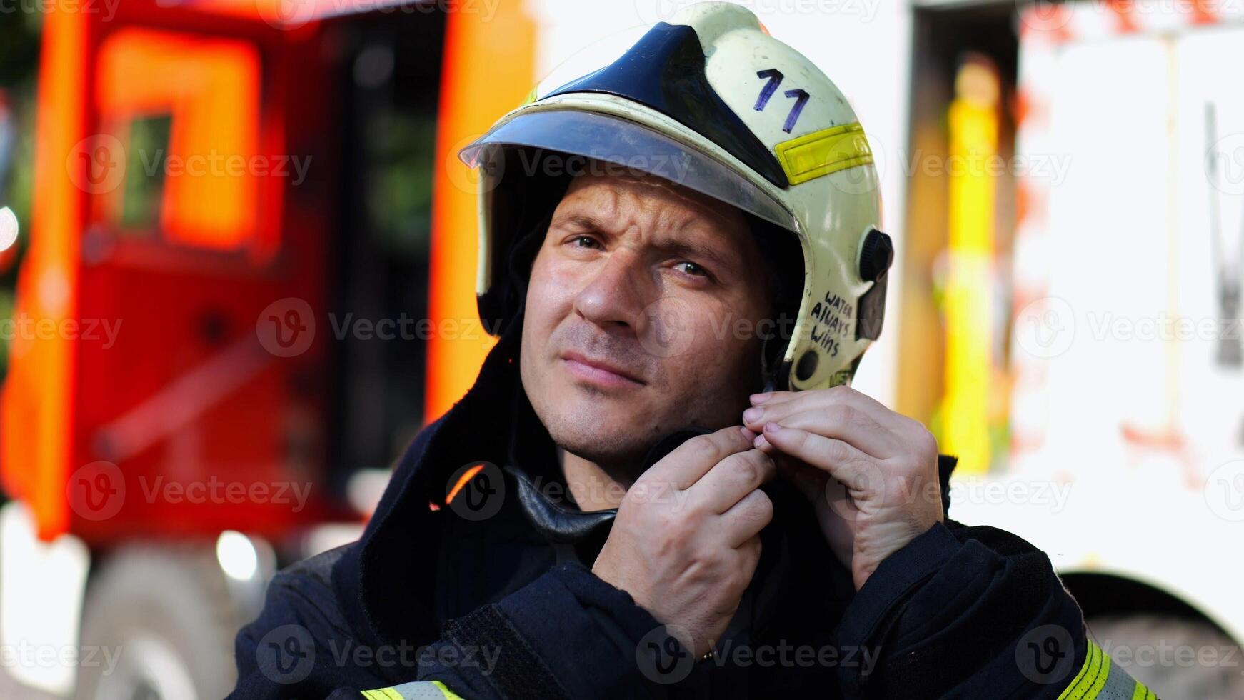 Young fireguard in protective uniform putting helmet against background of a big red truck. Male firefighter in full equipment near fire engine ready to work. Saving lives and heroic profession photo