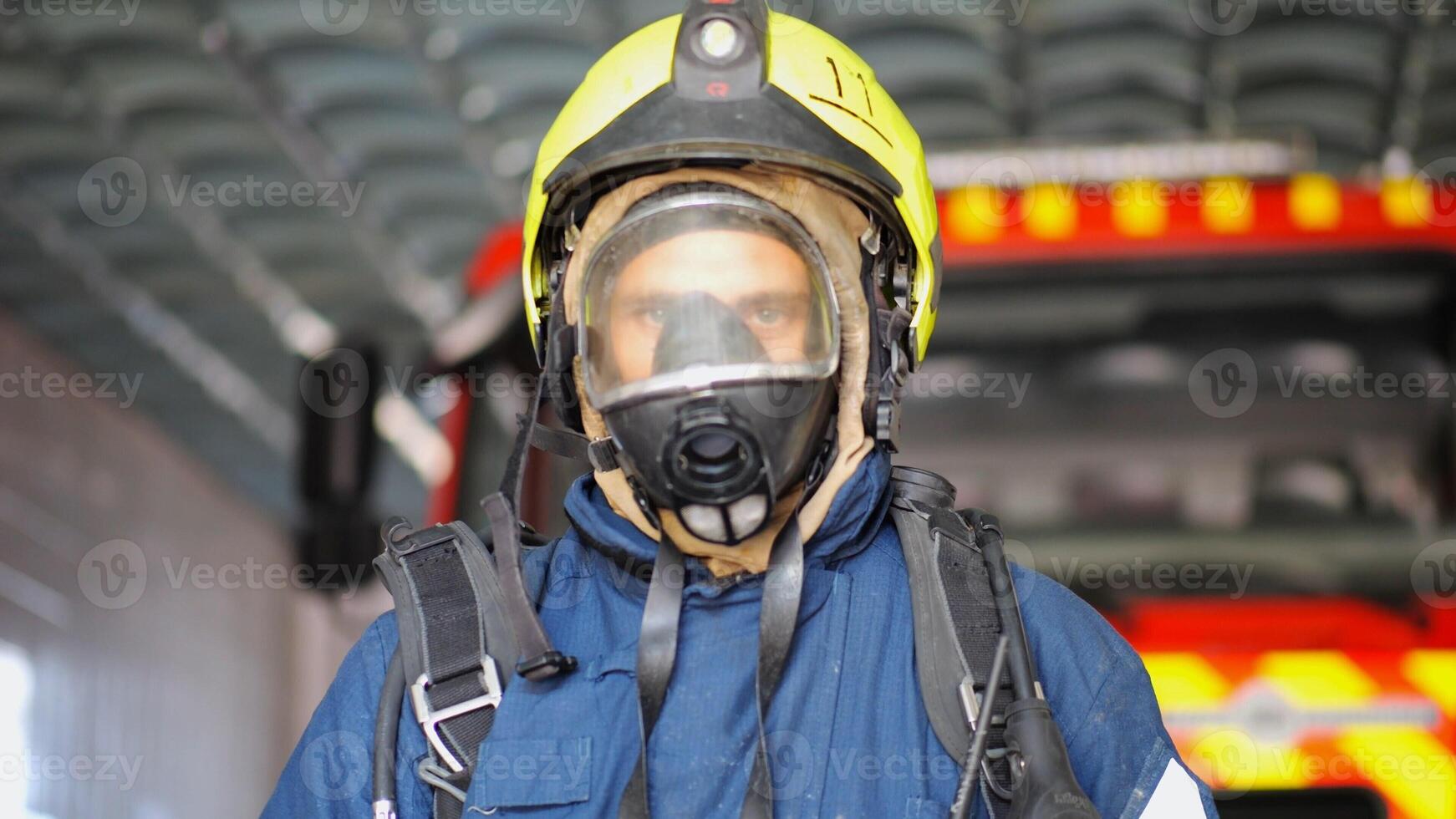 Portrait of male firefighter in protective helmet and uniform standing near a big red car at station. Young confident fireman in full equipment posing against the background of a fire engine. Close up photo