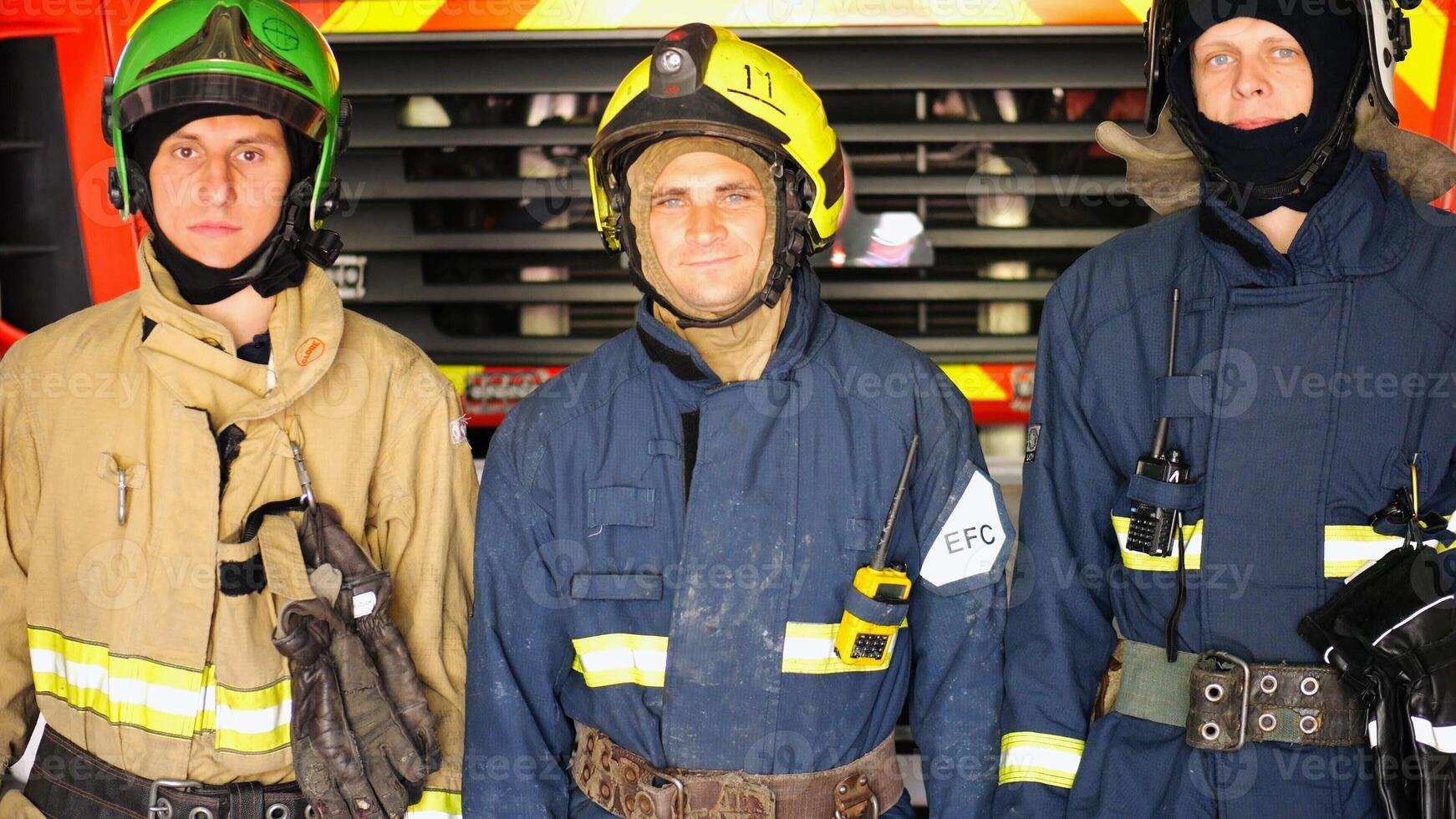 Portrait of male firefighters in helmets and protective uniforms standing near a big red car. Young confident firemen in full equipment posing against the background of a fire engine at station photo