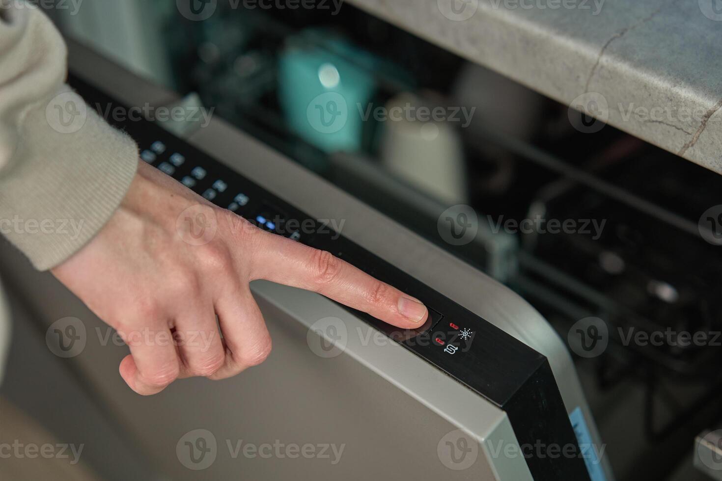 Hand pressing start button on dishwasher control panel with open door. Woman using modern home appliance, choosing automatic program on built in dishwasher for cleaning dirty dishes photo