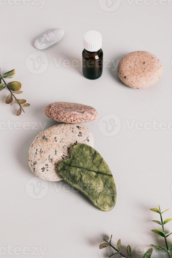 Jade gua sha massager on pebbles, essential oil on table vertical view photo