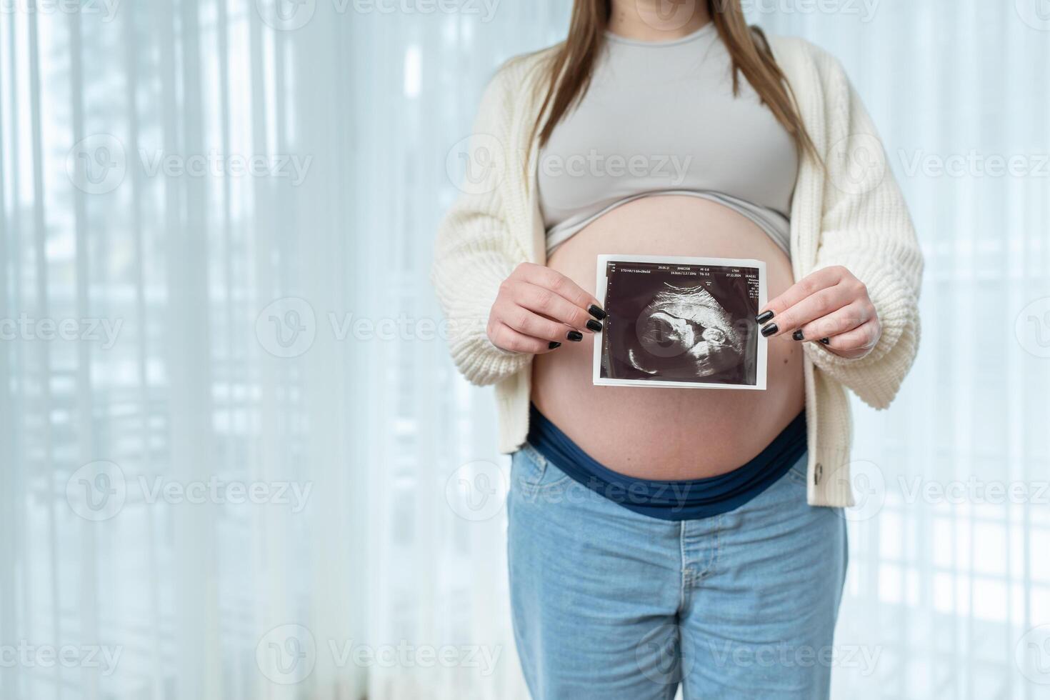 Young pregnant woman holding ultrasound scan photo of a fetus in front of her belly in third trimester at home with copy space for text. Concept of pregnancy, sonography. Maternity prenatal care
