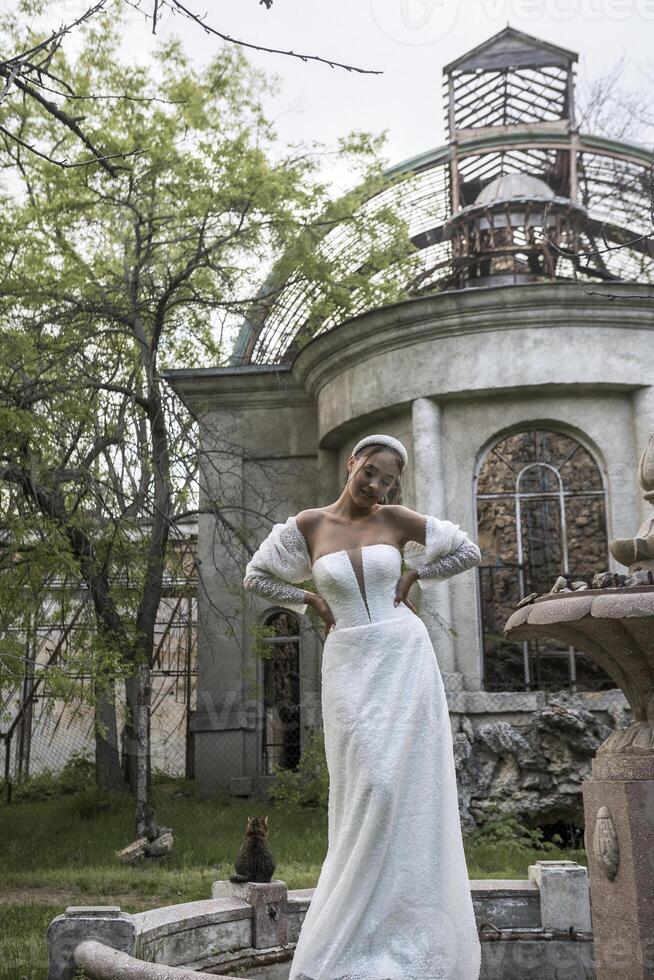 Bride with smooth hairstyle and white headband in hair against background of old building. Brunette girl in long white dress with open shoulders. Behind are ruins of fountain and 18th century building photo