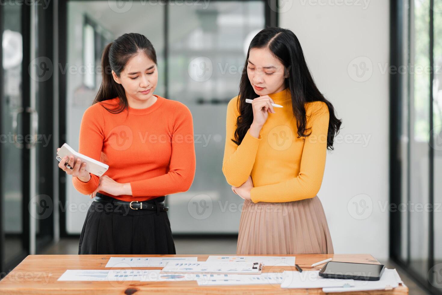 Two women in a modern office discuss a project, surrounded by documents and digital devices, showcasing teamwork and creativity. photo