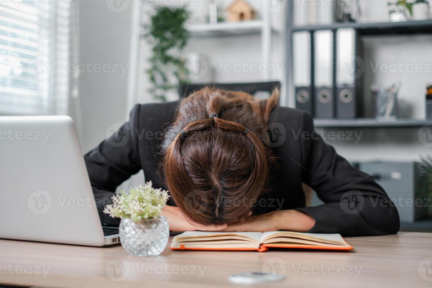 Tired businesswoman in office resting her head on a desk with a laptop and notebook, symbolizing stress and burnout. photo