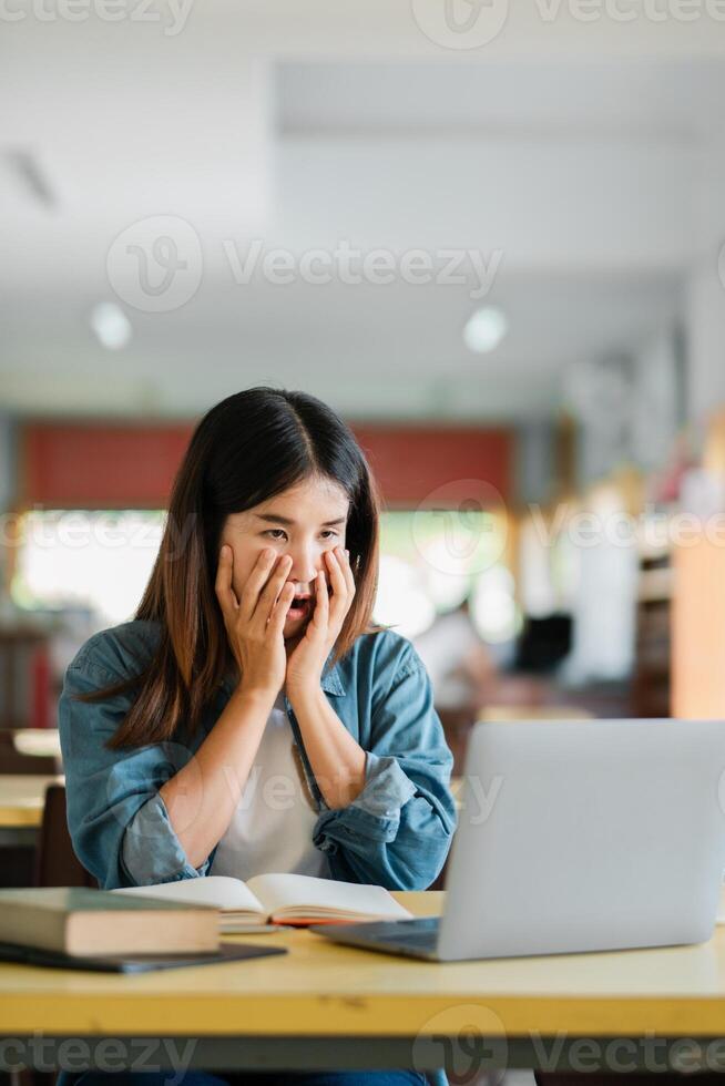 A young woman in a casual setting expresses surprise while using a laptop, surrounded by books in a cozy cafe environment. photo