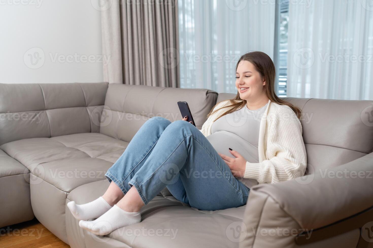 Smiling pregnant woman sitting on sofa at home using smartphone for online shopping and digital services with copy space. Concept of maternity, digital connection, technology and ecommerce convenience photo