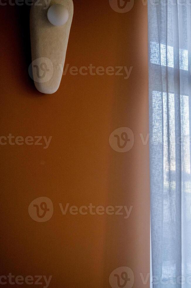 a wall of rich beige color with a lamp. next to a large window. the lamp is oval, it hangs on the wall with ceramic elements. photo