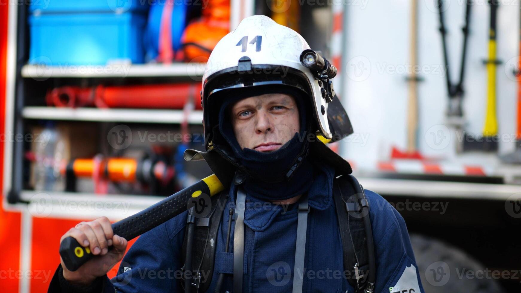 Portrait of confident male fireguard with axe standing near a fire engine. Young fireman with dirty face looking at camera in full equipment after fire against the background of big red truck photo