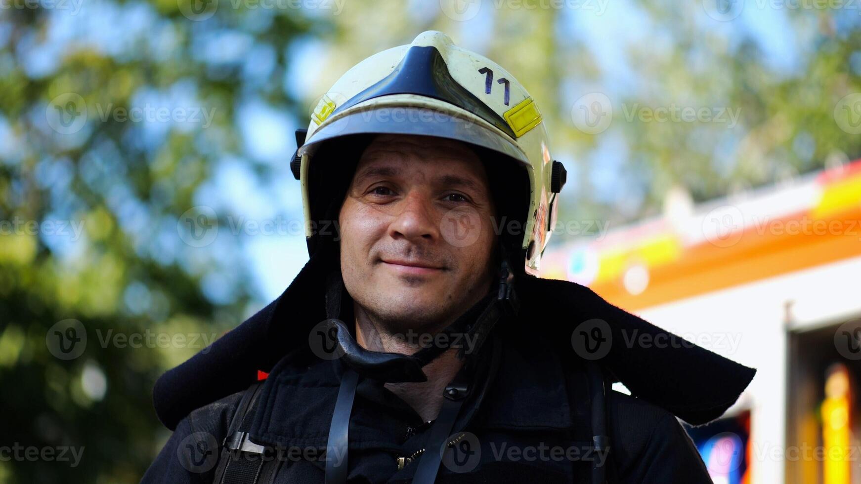 Portrait of young confident fireguard in protective uniform against background of a fire truck. Male firefighter in full equipment looking into camera with positive emotions near big red car. Close up photo