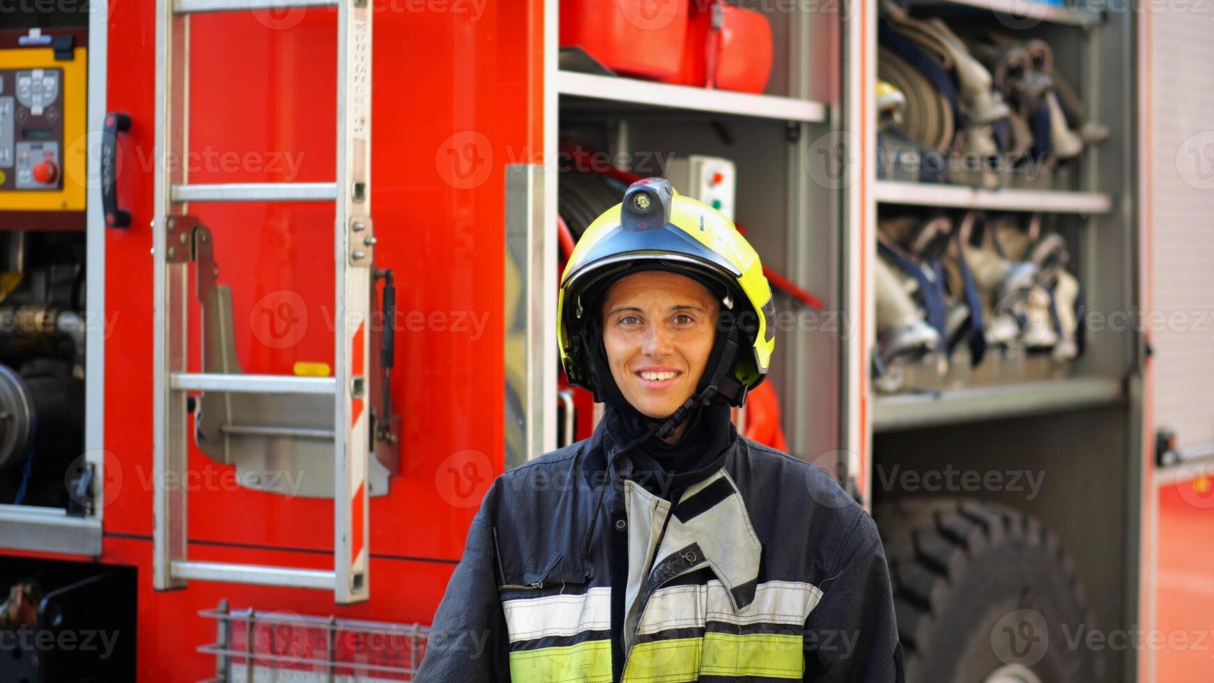 Portrait of happy female fireguard against the background of big red truck. Young firewoman in full equipment looking at camera with positive emotions near a fire engine. Concept of heroic profession photo