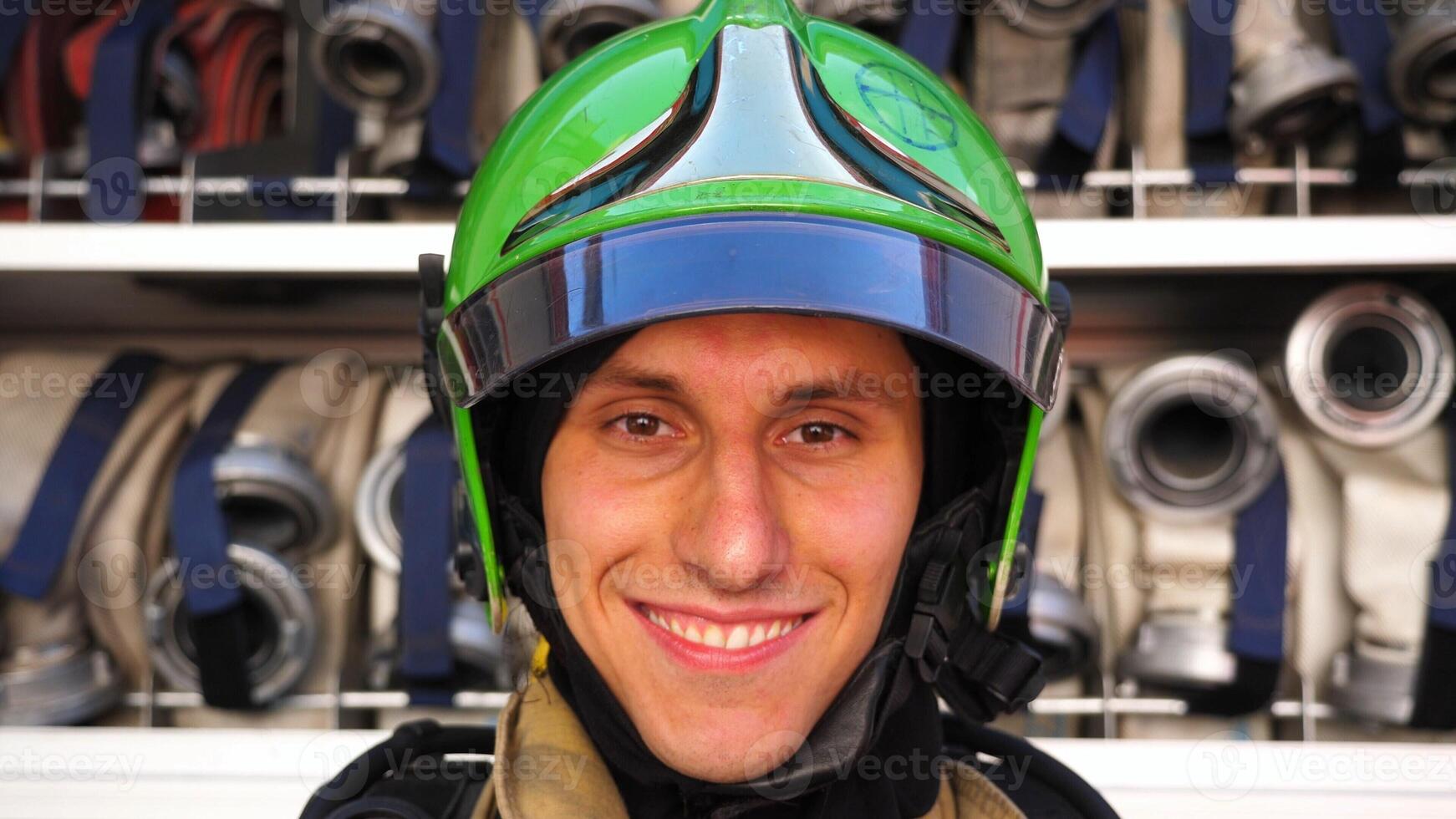 Young smiling fireman looking at camera in full equipment after fire against the background of big red truck. Portrait of happy male fireguard standing near a fire engine. Concept of heroic profession photo