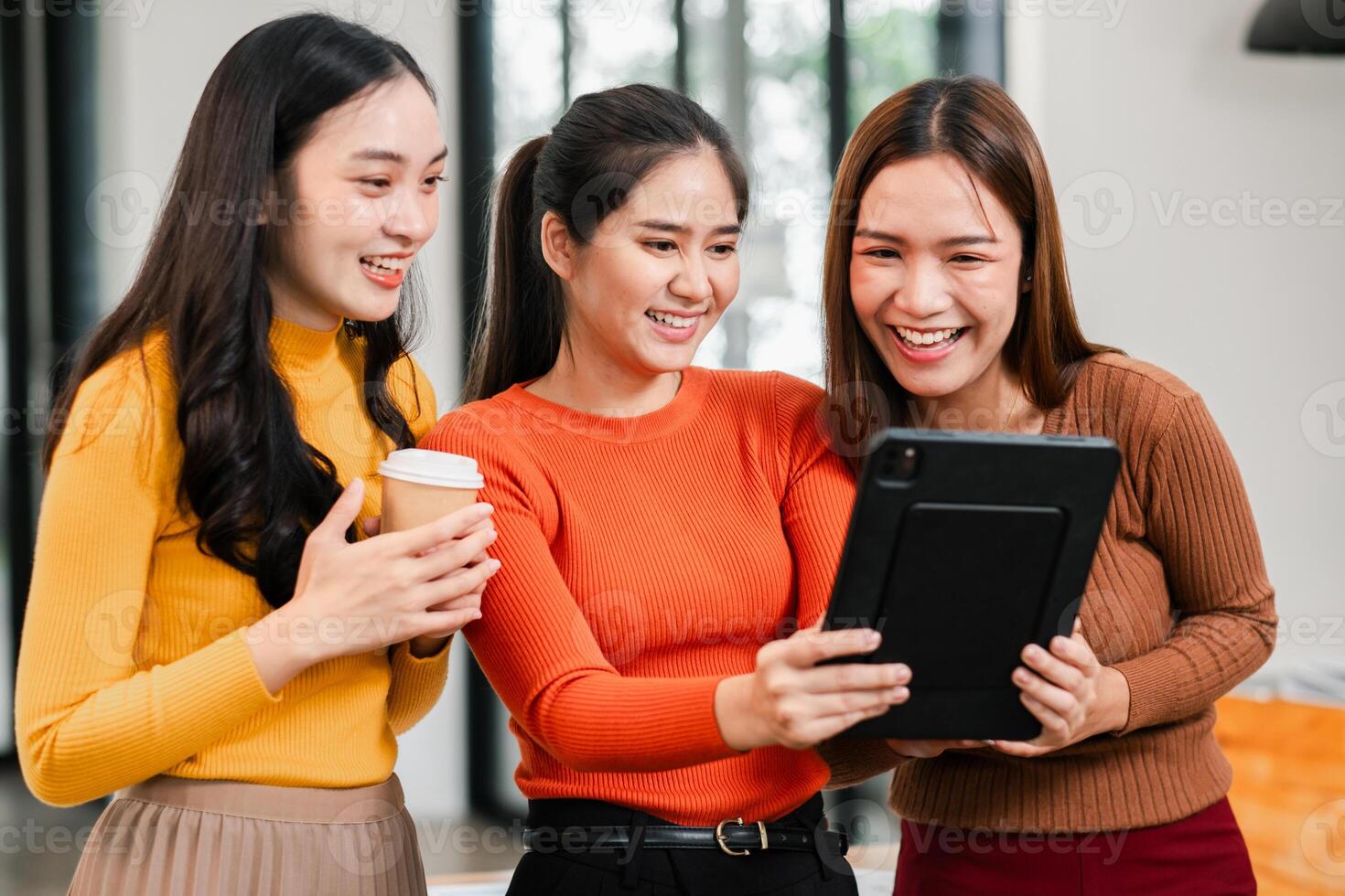 Three young women smiling and looking at a tablet together in a bright, modern office setting. photo