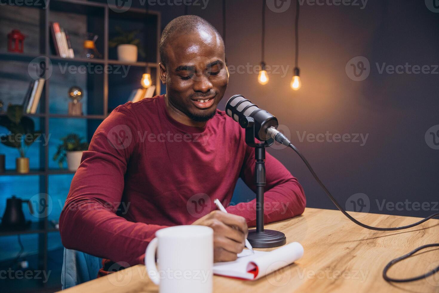 Young African American man recording a podcast episode, speaking into microphone and taking notes in notebook at home studio photo