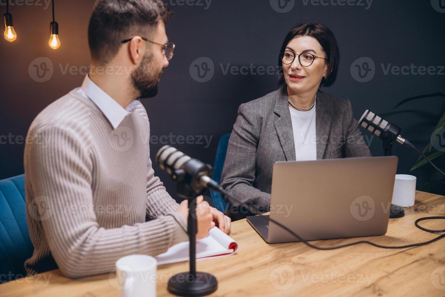 Two podcasters, a man and a woman, are recording a show in a professional studio, using microphones and a laptop photo
