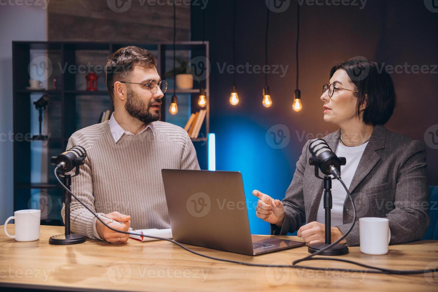 Two content creators recording their podcast using professional microphones and a laptop in a studio with soft lights photo