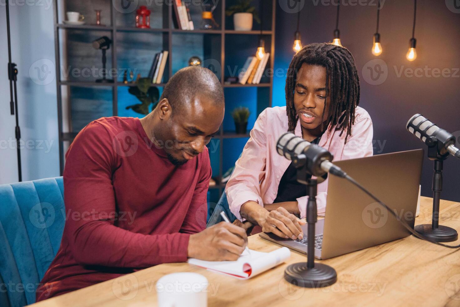 Two African American content creators recording a podcast episode in their home studio, using professional microphone and laptop photo