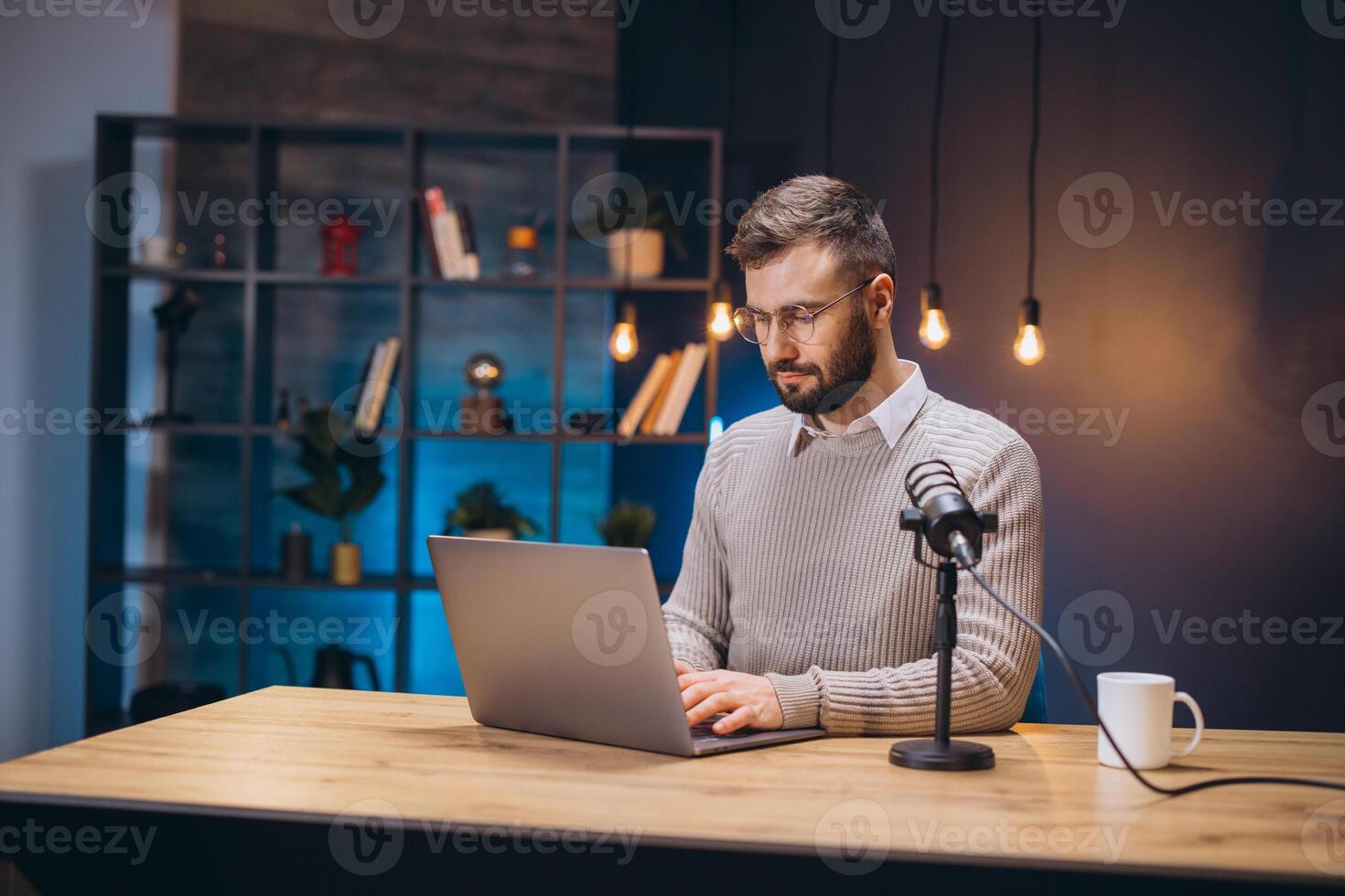Male podcaster speaking into microphone, working on laptop in intimate home recording space with soft lighting photo