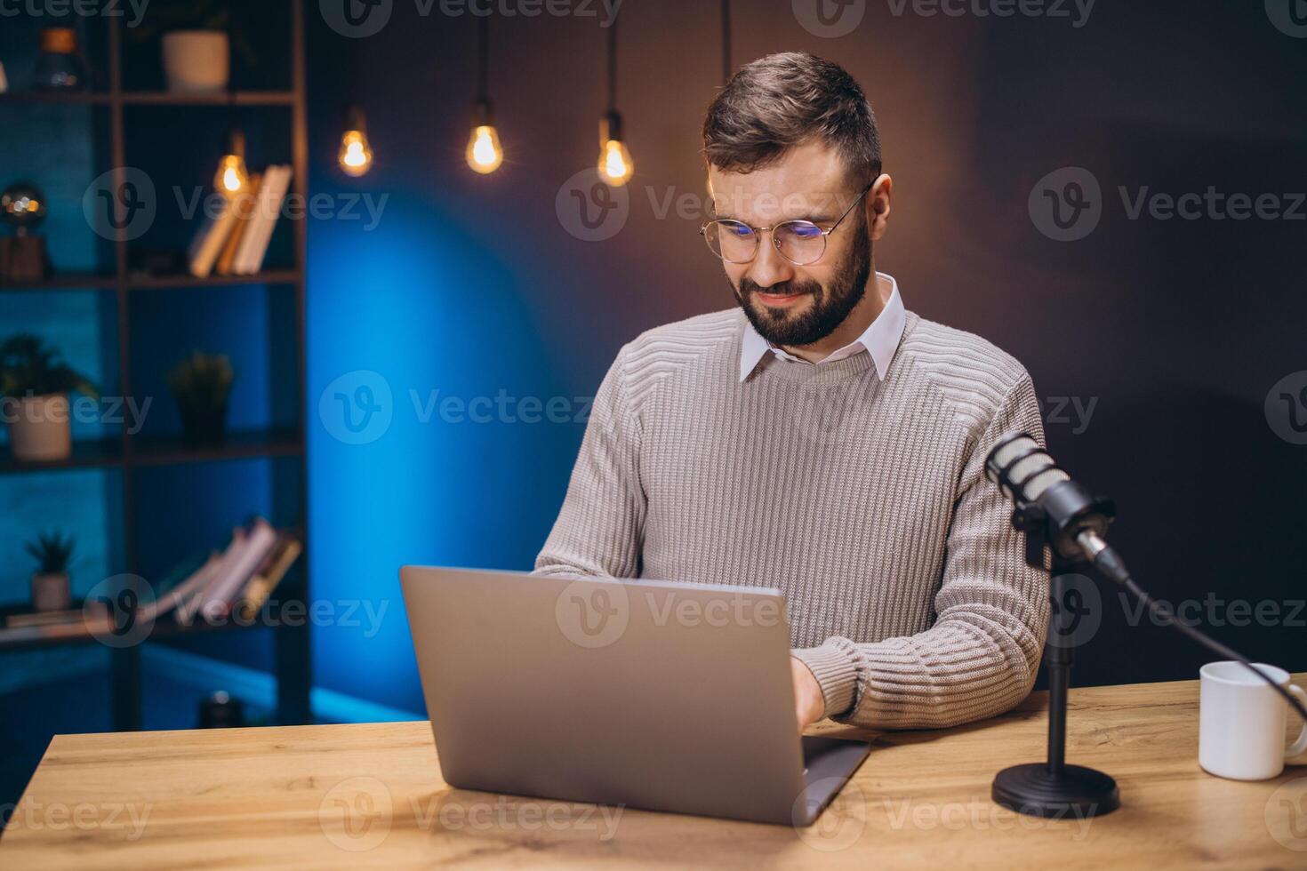 Young podcaster preparing content using laptop with microphone in home studio with blue light and bookcase photo