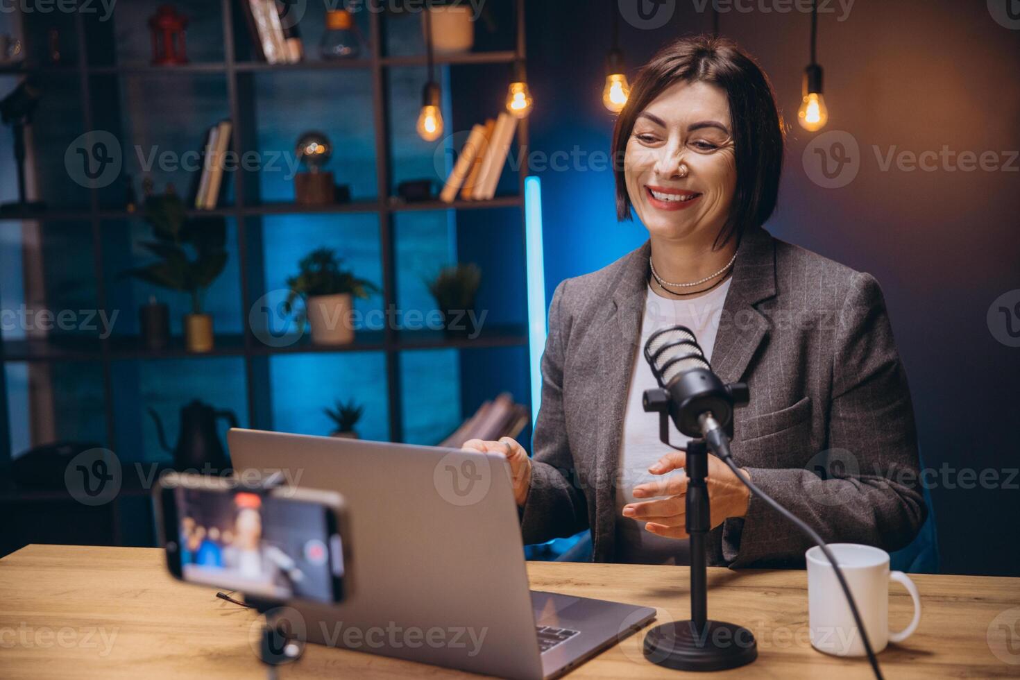 Smiling female podcaster recording audio content, speaking into microphone while using laptop and smartphone in home workspace photo