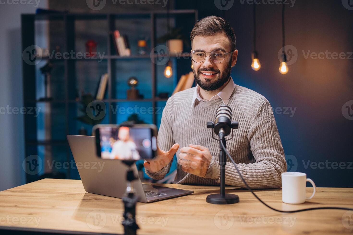 Male podcaster speaking into mic, recording digital content with smartphone in home workspace, wearing casual attire photo