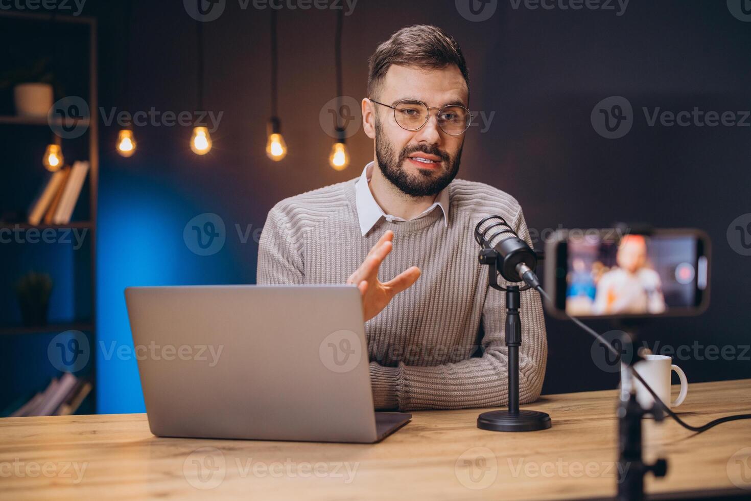 Young man recording a podcast using a professional microphone and laptop, talking and gesturing while looking at the camera photo