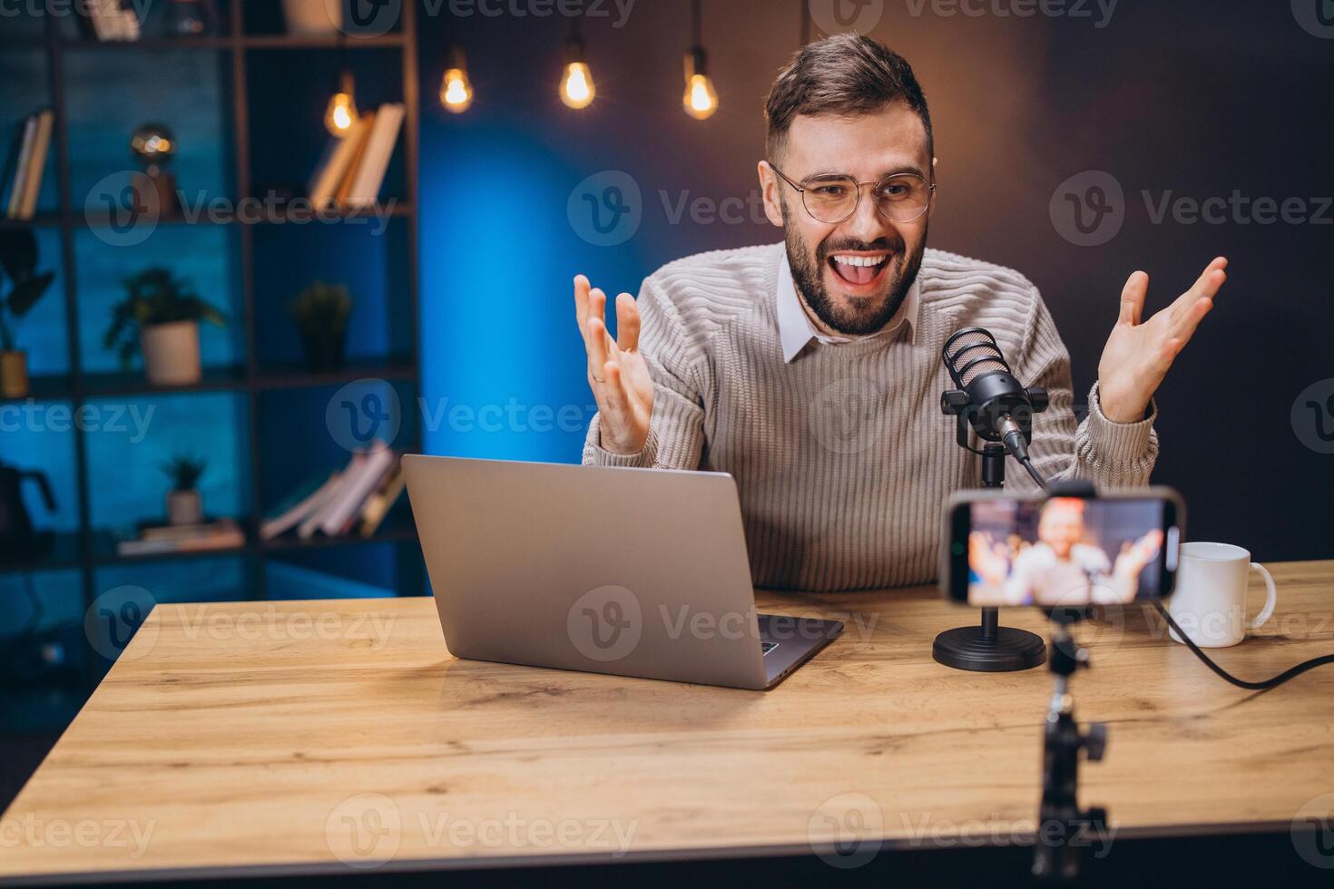Happy male influencer gesturing while recording a podcast in his home studio using professional microphone, laptop and smartphone photo