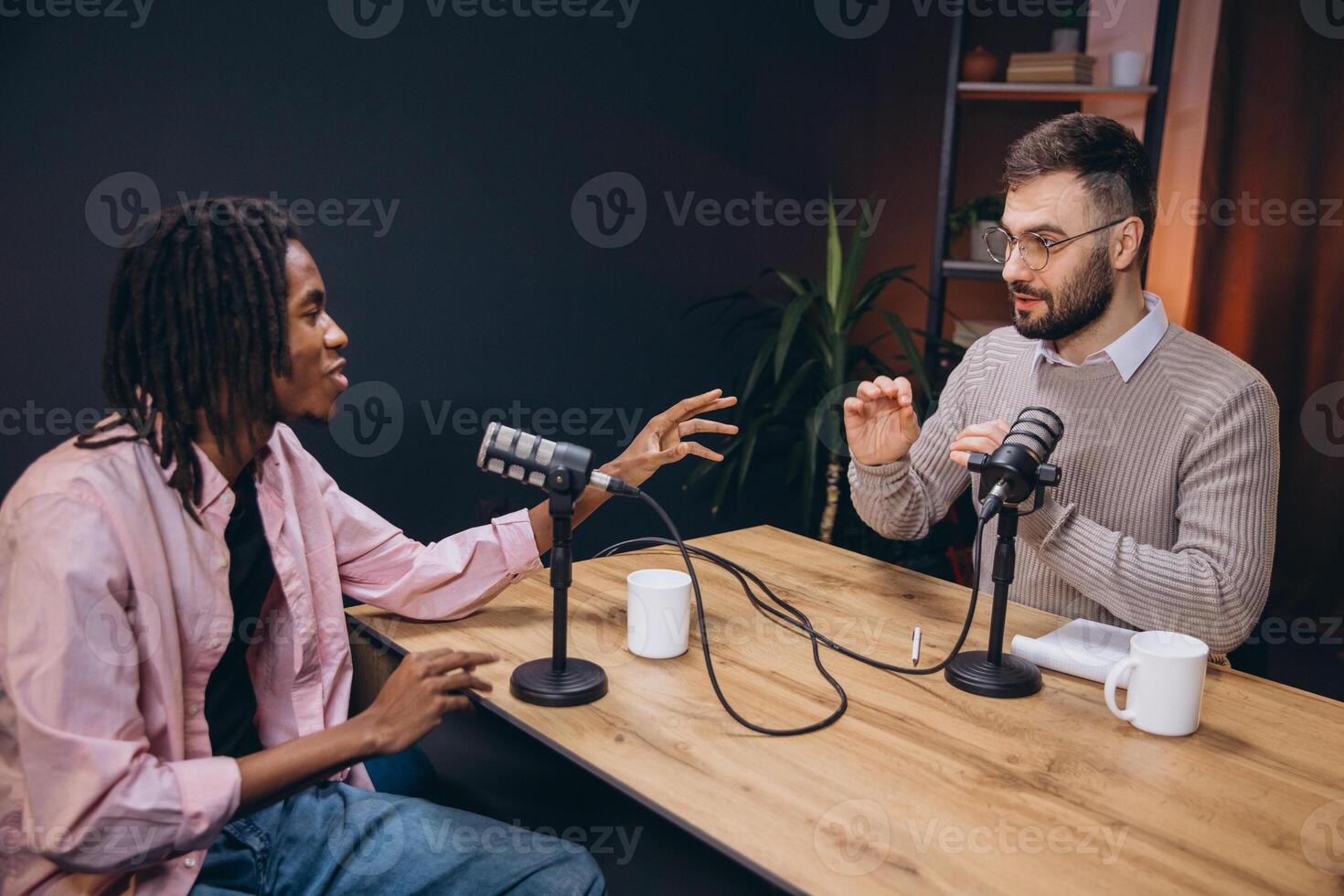 Two male podcasters sitting at a wooden table talking and gesturing during a recording session in a professional studio photo