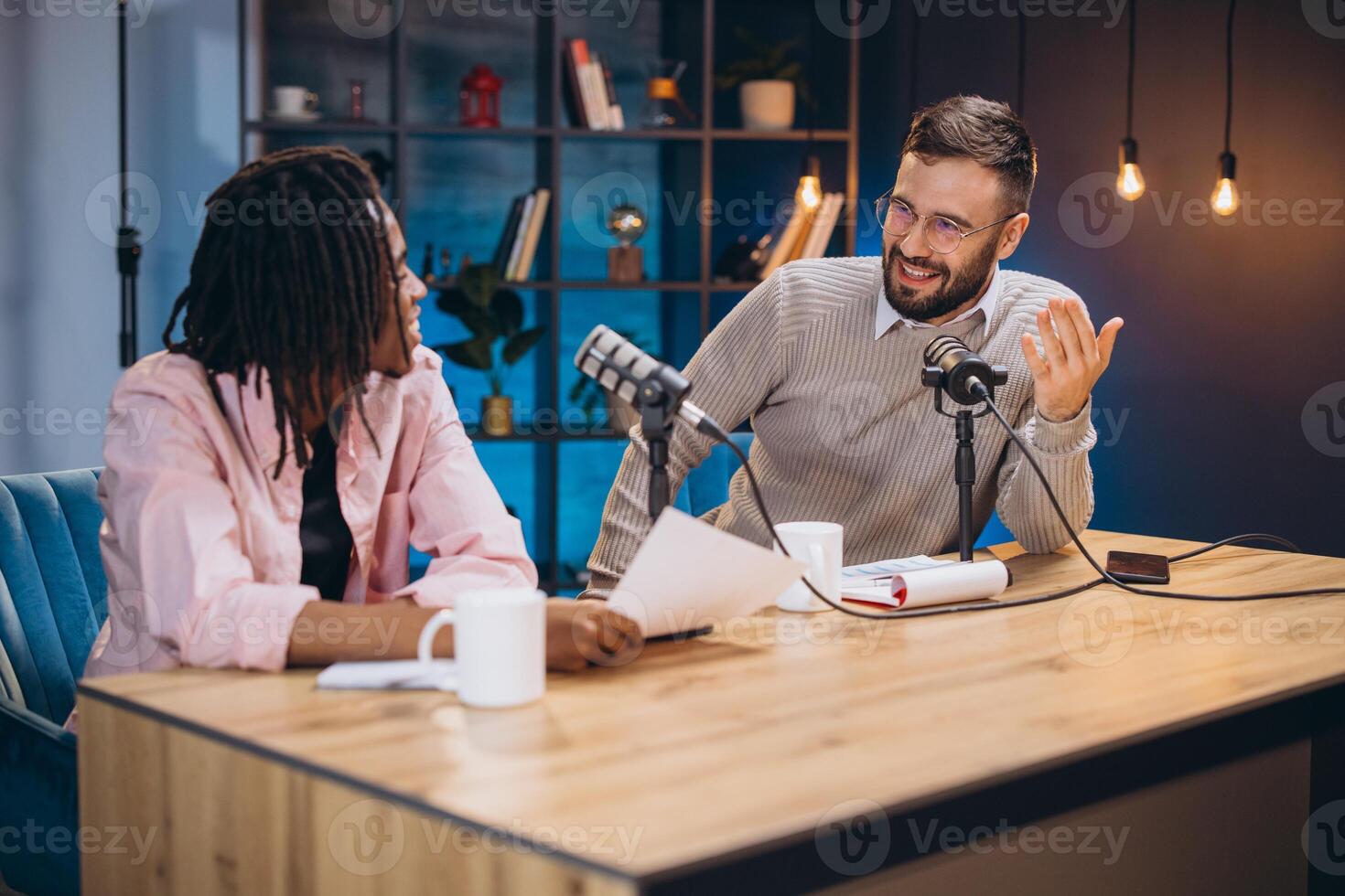 Podcasters recording episode, discussing content with microphones, notes spread across workspace, capturing dynamic conversation photo