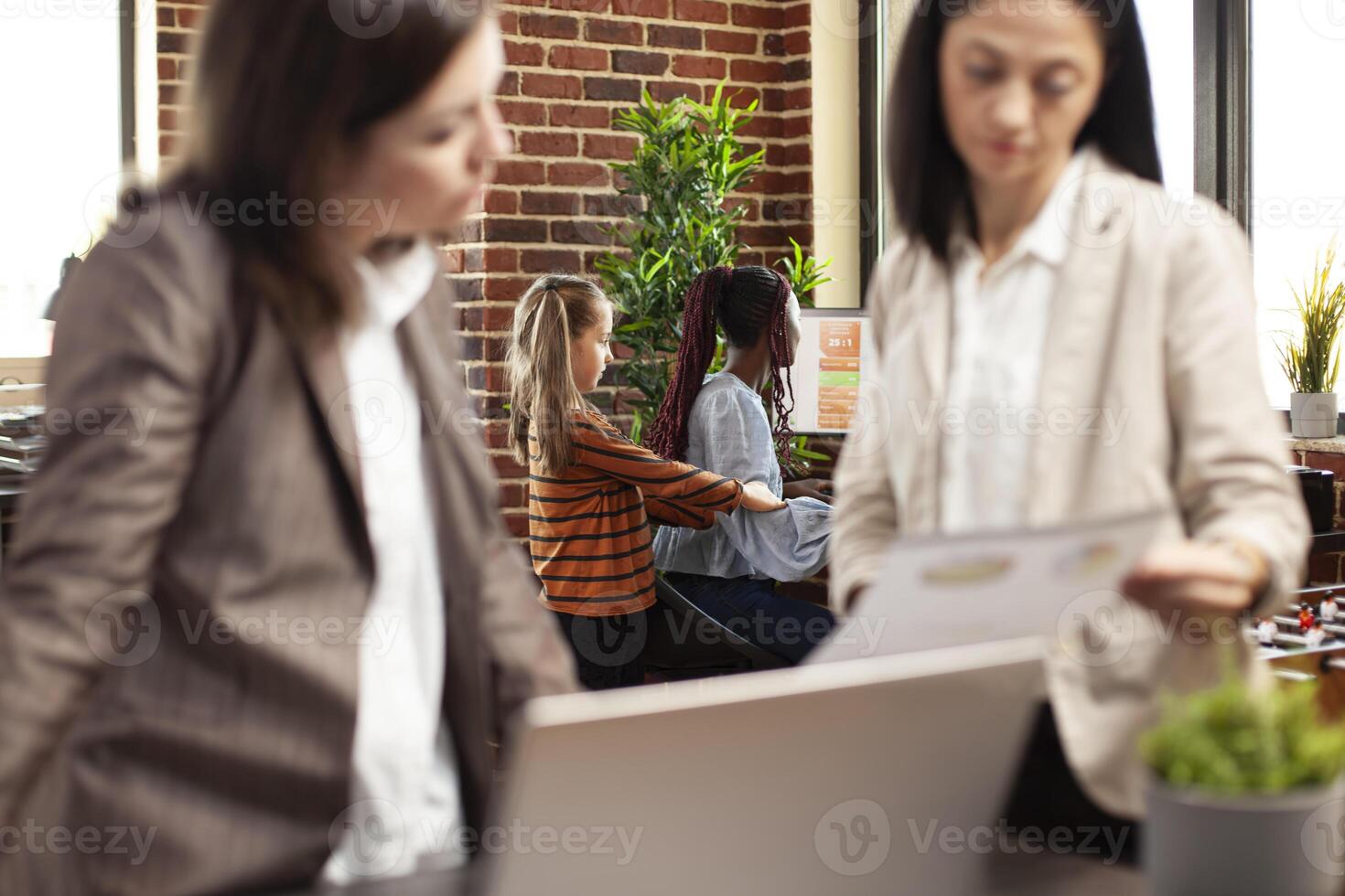 Bored girl interrupts african american employee trying to work on computer at startup office. Colleagues review documents as child distracts black woman researching financial charts on desktop pc. photo