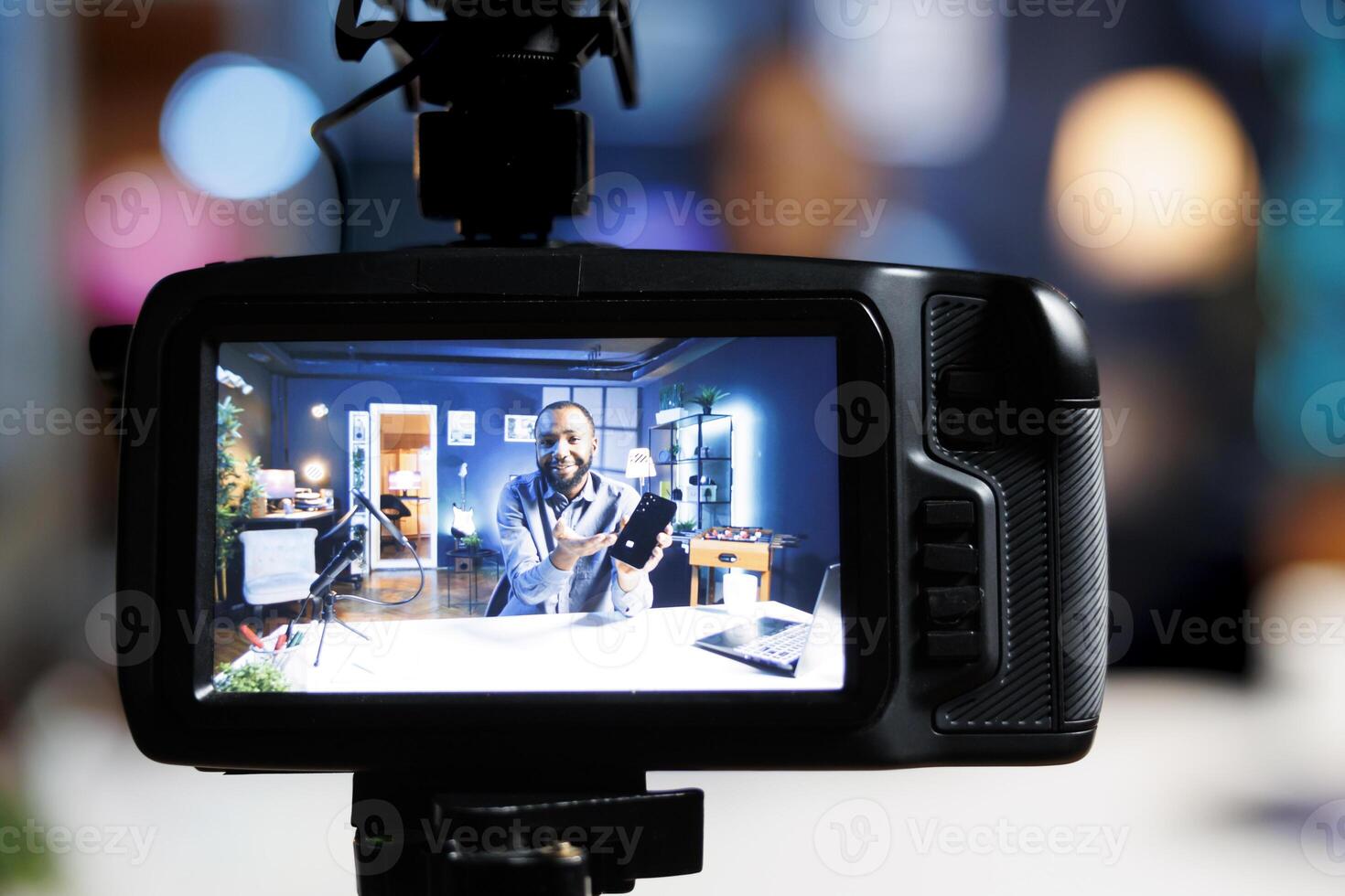 Focus on professional production camera used for capturing footage of tech engineer in blurry background filming newly launched smartphone review for online audience, showcasing mobile phone photo