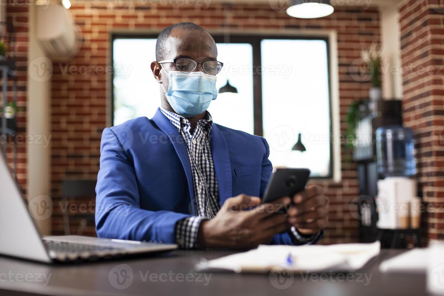 Portrait of businessman with face mask texting on his mobile device in brick wall office. African american manager taking a break at work, using smartphone for chatting and checking social media. photo