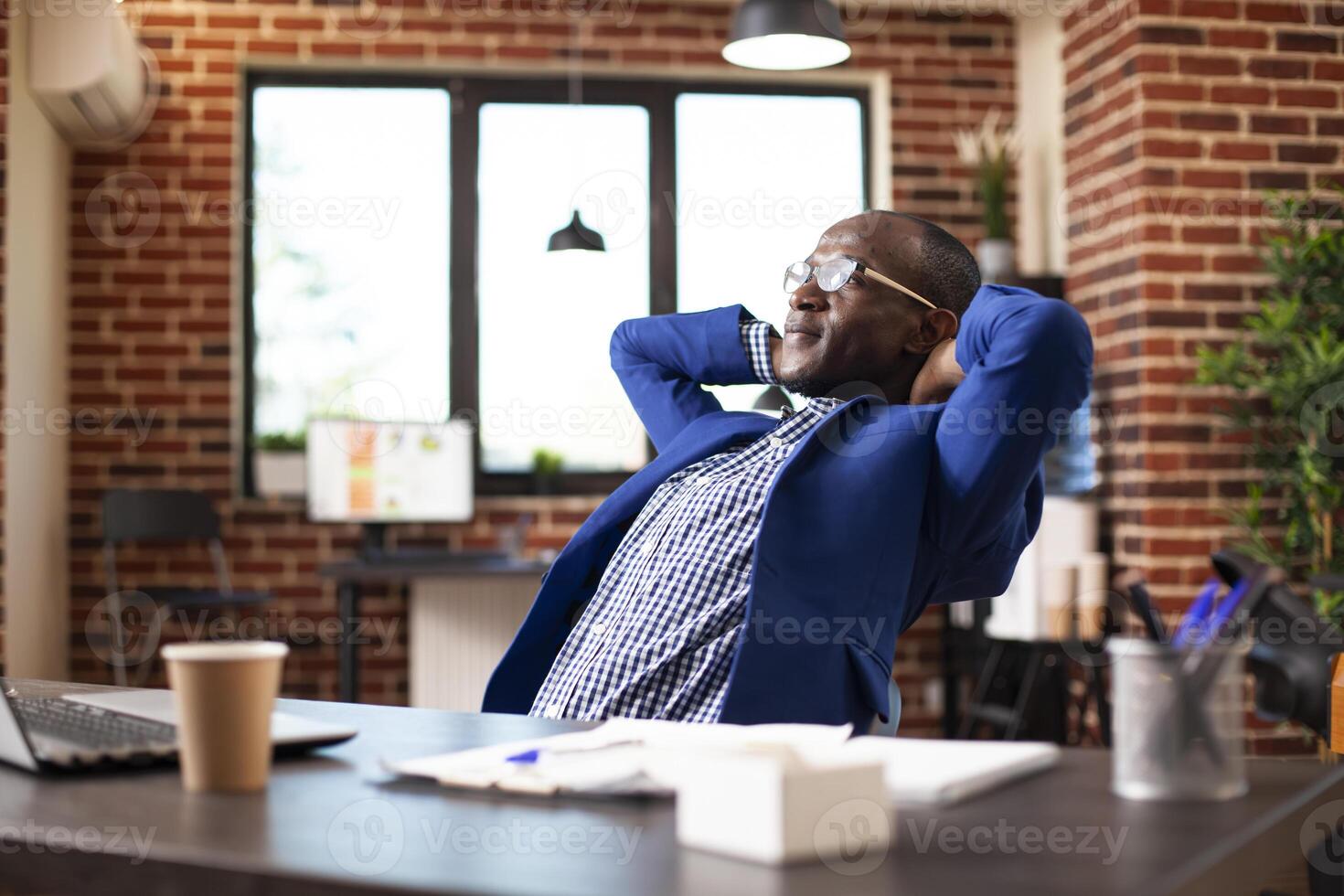 Male manager seated at desk, leaning on chair with his hands behind head in brick wall room. African american man in business clothing taking a break by relaxing at his table in startup office. photo
