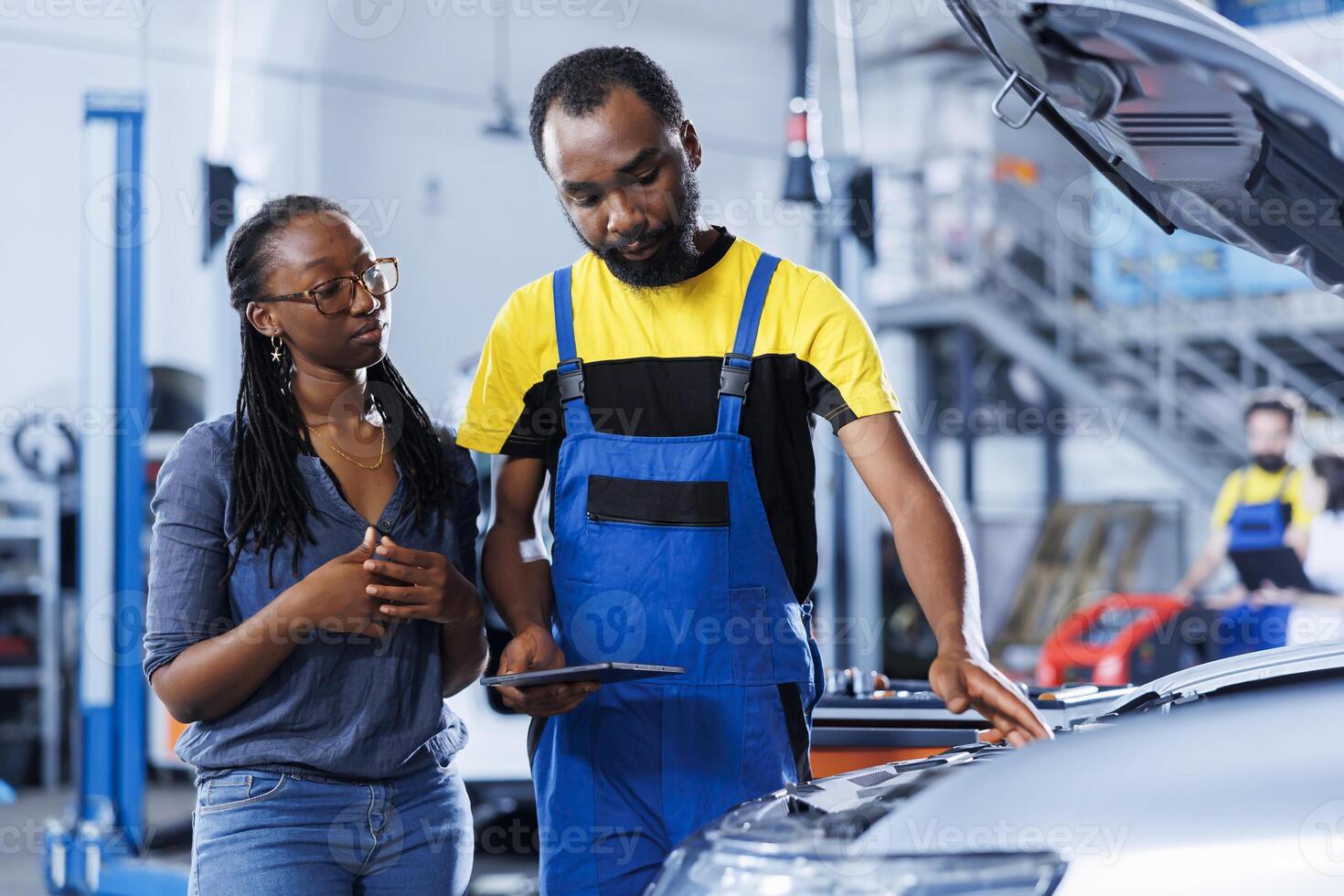 BIPOC mechanic in car service uses tablet to calculate invoice for client after repairing motor. African american employee in garage with customer calculating final costs after servicing vehicle photo