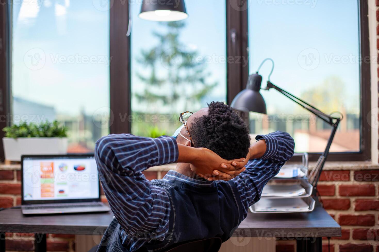 African american manager sits at desk, leaning back in his chair with hands behind head, enjoying quiet break in startup office. Black male employee looking out the window and reflecting on workday. photo