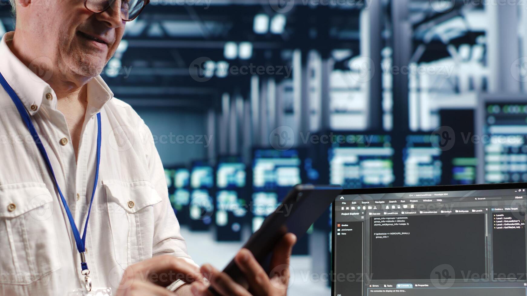 Senior technician in data center, solving tasks on tablet while running scripts on laptop terminal. Licensed serviceman amidst servers, accessing databases and manipulating lines of code photo