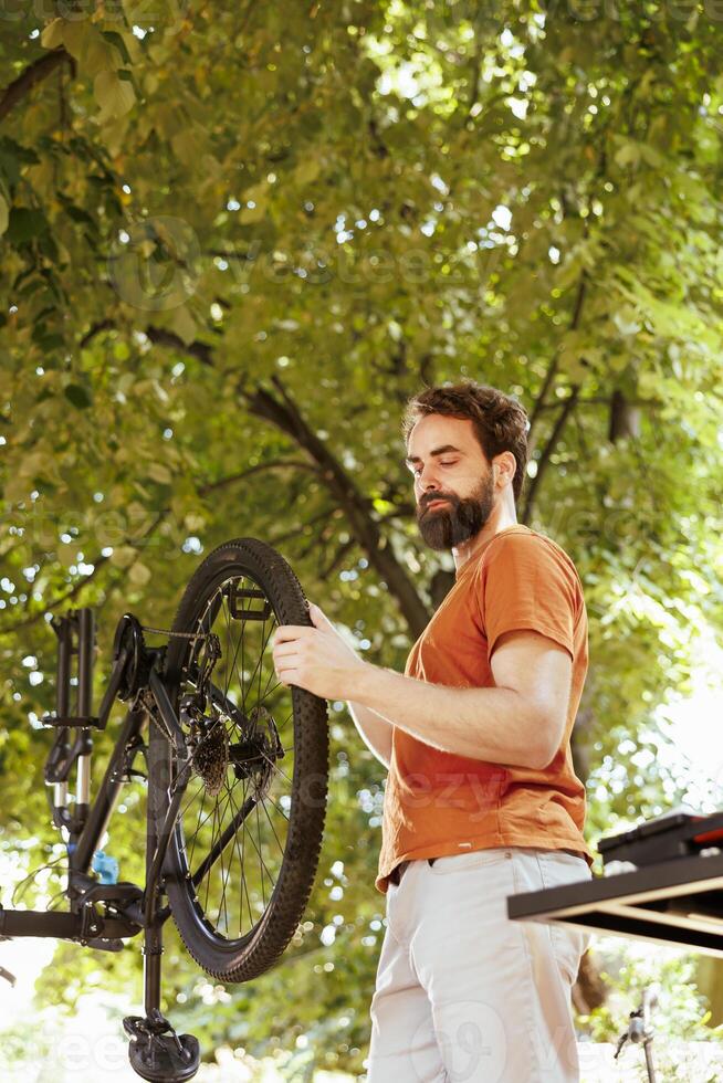 Committed sports enthusiast man working outside in summer on modern bicycle for leisure cycling. Active caucasian male cyclist thoroughly inspecting for damages on bike to fix and repair as annual maintenance. photo